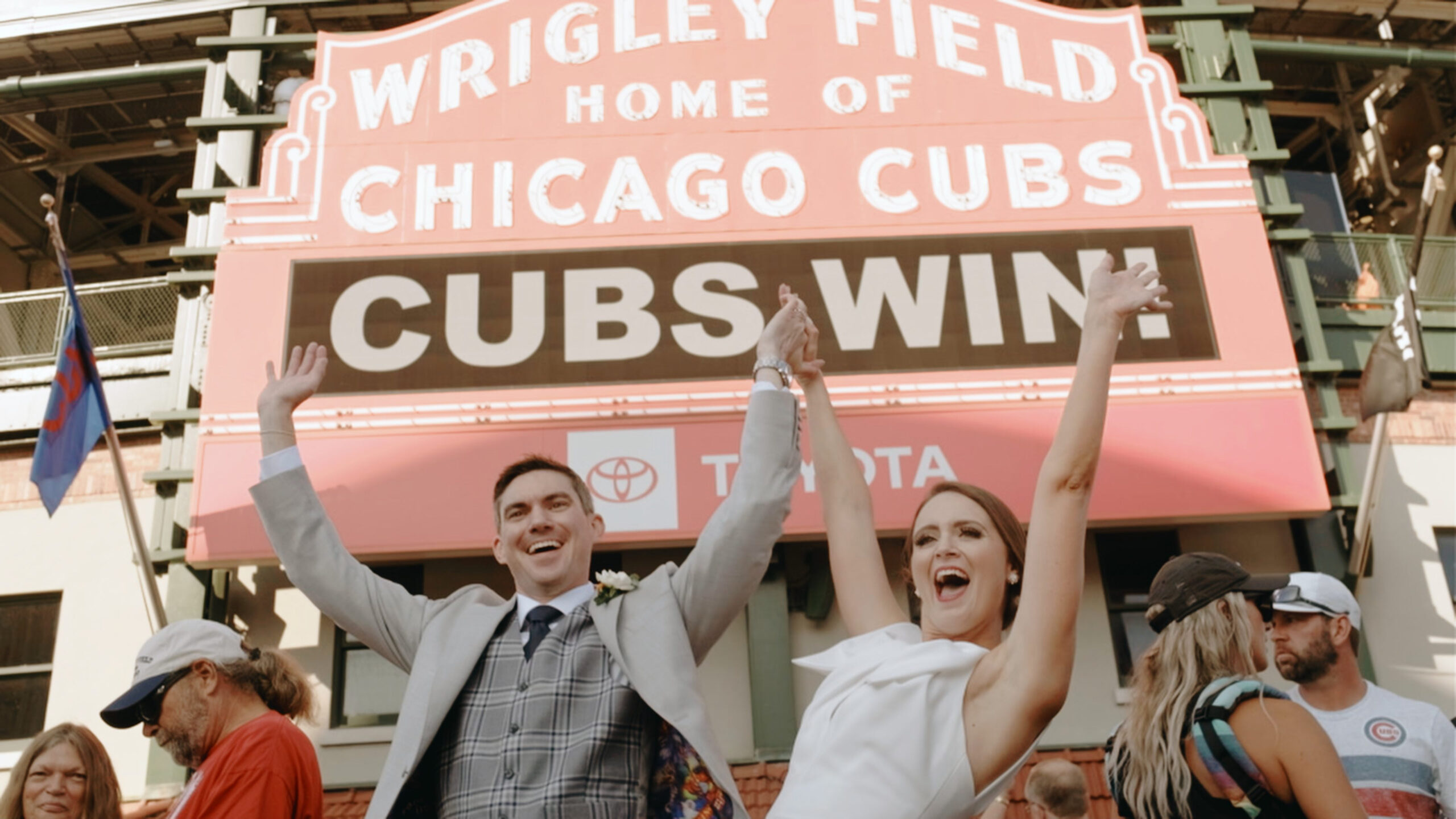 a bride and groom hold hands and toss them in the air in celebration in front of the wrigley field sign that reads "Cubs Win!" - a biagio events wedding